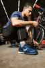 Preview: A man is kneeling in a workshop working on a bicycle. He is wearing sporty clothing and blue shoes. The background displays tools and bicycle parts.