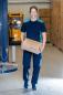 Preview: In the picture, there is a woman in blue work attire carrying a cardboard box. She is smiling and is in a warehouse with many boxes in the background.