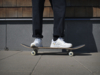 Preview: In the picture, a person is standing on a skateboard. They are wearing white sneakers and dark, baggy pants. The background consists of wooden panels and a gray wall.