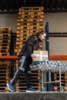 Preview: A man in work clothes is lifting a box in a warehouse. In the background, wooden pallets are stacked. He is standing next to a cart with several boxes.