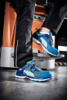 Preview: The image shows two feet in stylish blue sports shoes standing on a lift platform. The background is industrialized, with orange and gray elements.