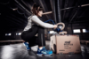 Preview: A person in sportswear is kneeling in front of a cardboard box on the floor. They are using a machine to work on something with a hose. The background is a warehouse setting with wooden pallets.