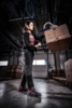 Preview: A woman is lifting a box in a warehouse. She is wearing work clothes and safety shoes. In the background, wooden pallets and a modern warehouse environment are visible.