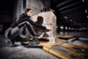 Preview: A person in athletic clothing bends down to secure a box on a pallet. The background shows a warehouse with large windows and industrial lighting.