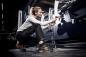 Preview: A woman, dressed in a gray sweatshirt and work pants, kneels next to a machine in a workshop. She is holding a tool and working intently. The setting is dim, with a hint of industrial ambiance.