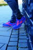 Preview: The image shows a shoe in vibrant blue with neon orange accents. A person stands on a paved path, partially obscured by jeans. The shoe has a sporty shape.