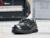 Preview: The image shows a black safety shoe with laces. It features a sturdy, padded cap and a non-slip sole. In the background are blurred industrial elements.