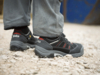 Preview: The image shows the feet of a person in sturdy, black work shoes on a rocky surface. The pant legs are wide and gray, and there are colored accents on the shoes.