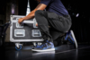 Preview: A man in a blue shirt and black pants is kneeling and opening a large, sturdy suitcase with wheels. He is wearing athletic shoes. Behind him is a stage.