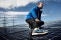 Preview: A man in a blue jacket and athletic shoes is squatting on a solar panel. In the background, power lines and a clear sky are visible. The scene exudes activity and technology.