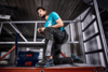 Preview: A young man is kneeling on a platform in work clothes. He is wearing a turquoise T-shirt and black pants with gray details. In the background, steel beams and a ladder are visible.