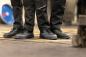 Preview: In the picture, there are three pairs of feet in black work shoes. The wearers are in black pants. The floor appears industrialized, with yellow markings.