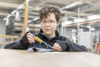 Preview: A boy with curly brown hair and a black jacket is sitting at a table. He is holding a screwdriver and looking focused. In the background, shelves and workshop tools can be seen.