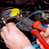 Preview: A hand holds a wire cutter with red and blue handles while working on a blue cable leading to a car component. In the background, a yellow container is visible.