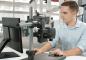 Preview: A young man is sitting at a table in front of a monitor. In front of him is a microscope with a large lens, mounted on a sturdy stand. He is focusing on his work.