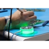 Preview: A section featuring a hand holding a soldering tip, while a green, glowing, circular device is on the table. Cables are connected to it, and circuits are visible in the background.