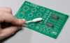 Preview: The image shows a green circuit board with various circuits. A hand is holding a cotton swab used to clean the board.