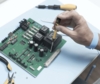 Preview: A hand holds a screwdriver and works on a green circuit board with various components. Next to the board, pliers are lying on a table with a blue surface.