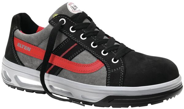 The shoe is a sporty sneaker with a gray and black surface. It features red accents, a padded tongue, and a textured sole for good grip. The laces are black.