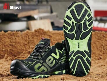 The image shows a black shoe with green accents and the brand "Sievi". The shoe has a profiled sole and is lying on sandy ground.