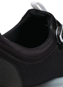 The image shows the upper part of a black sports shoe. The shoe has a soft, padded material and a round cutout. A closure system is depicted at the top.