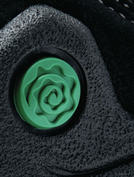 The image shows a black shoe with a round, green detail in the shape of a curled rose. The surface appears textured and slightly rough.