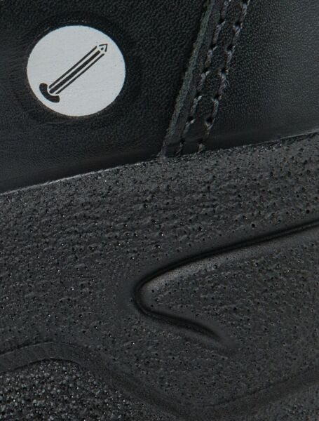 The image shows a part of a black shoe made from textured material. On the surface, there is a small round symbol featuring a pencil.