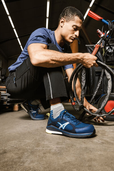 A man is kneeling in a workshop working on a bicycle. He is wearing sporty clothing and blue shoes. The background displays tools and bicycle parts.