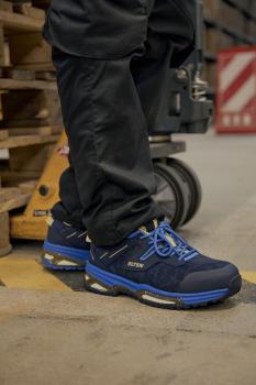 The image shows a standing man in dark pants wearing blue and black safety shoes. The background is a warehouse with wooden pallets.