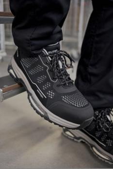 The image shows a black athletic shoe with a breathable upper and a thick, grippy sole. It is positioned on a metal step, surrounded by a neutral background.