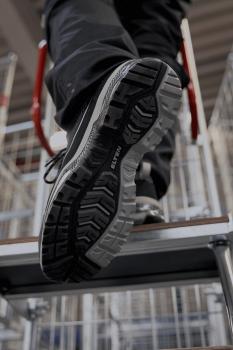 The image shows the underside of a sports shoe standing on a step. The shoe has a black and gray treaded sole and is part of a person standing at the top of a staircase.