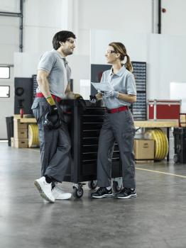 The image shows two people in work clothes standing at a tool cart. They appear friendly and are conversing. In the background, tools and boxes are visible.