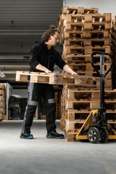 A person in dark work clothes is lifting a wooden pallet. In the background, many stacked wooden pallets can be seen. The floor is gray and the surroundings appear industrial.