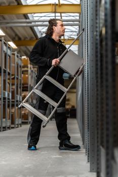 A man in black work clothes stands in a warehouse holding an aluminum ladder. He looks up at the shelves to reach something. The environment appears industrial and well-organized.