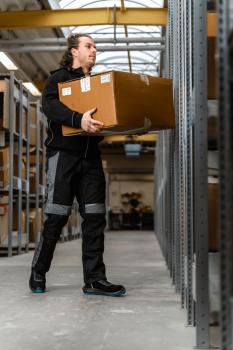 A man is carrying a large box in a warehouse. He is wearing black work clothes and is standing between shelves. The room has a high ceiling and is well-lit.