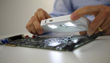 A person holds a light-emitting magnifying glass over a green circuit board. The hands are visible, and the device displays components such as chips and holes for connections.