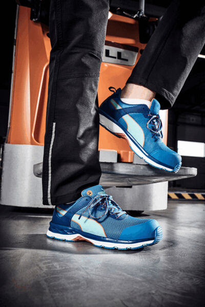 The image shows two feet in stylish blue sports shoes standing on a lift platform. The background is industrialized, with orange and gray elements.