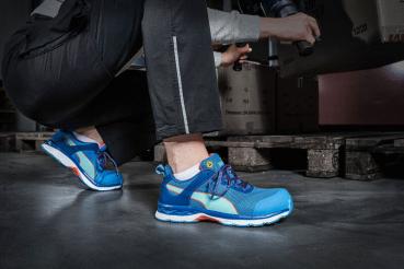 A person is kneeling on the ground in an industrial setting. They are wearing bright blue athletic shoes with white soles. The background features several pallets and devices.