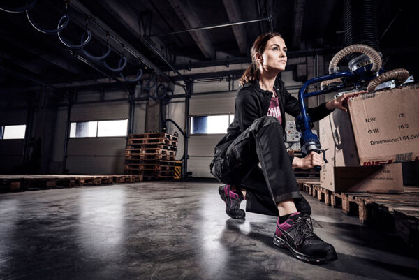 A woman is kneeling in a modern warehouse. She is wearing athletic clothing and has a determined expression on her face. Around her are wooden pallets and boxes stacked in various places.