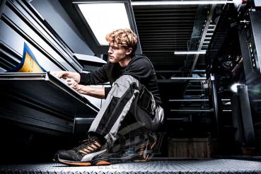 A young man in work clothes is sitting on a metal floor studying documents. He has curly, light hair and is wearing shoes with orange accents. Behind him is an industrial space.