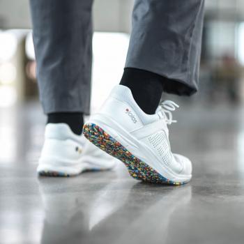 The image shows a foot in a white sneaker with a colorful, textured rim on the sole. The wearer is wearing gray pants and black socks. The surroundings appear modern and bright.