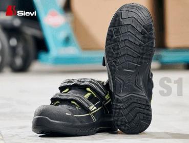 The image shows a black safety shoe with two straps and a non-slip, textured sole. It has a sturdy, sporty shape and a green accent color.