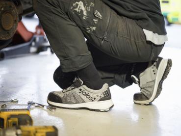 A person is sitting on the floor, wearing black work pants and gray athletic shoes. Tools are scattered on the floor, and the surroundings resemble a workshop.