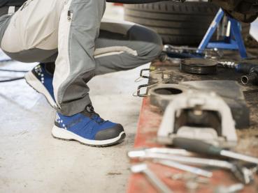 The image shows a person in work clothes, sitting at knee height in front of a car. They are wearing blue shoes and working with tools on a workbench, surrounded by car parts.