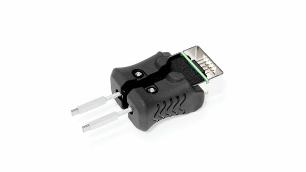 The image shows a black plug with a rectangular connection on top and two metallic pins protruding from the sides. The surface is ribbed and there are small screws.