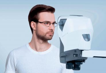 A young man with glasses stands next to a modern, white eye examination device. The background is in soft shades of blue. He is wearing a plain, white T-shirt.