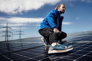 A man in a blue jacket and athletic shoes is squatting on a solar panel. In the background, power lines and a clear sky are visible. The scene exudes activity and technology.