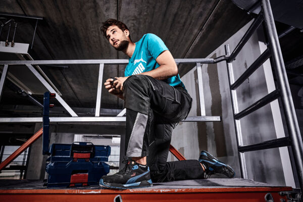 A young man is kneeling on a platform in work clothes. He is wearing a turquoise T-shirt and black pants with gray details. In the background, steel beams and a ladder are visible.