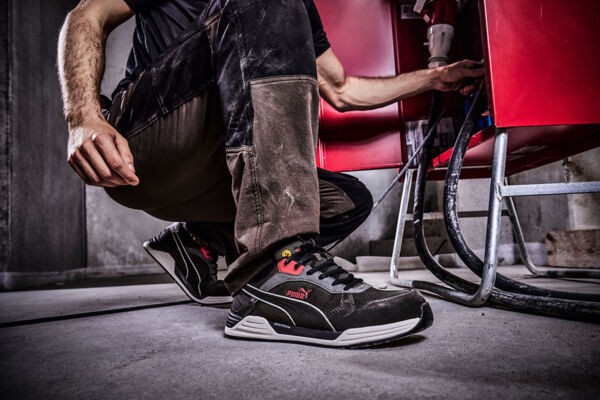 A man is kneeling on the ground in work clothes with dirty pants and safety shoes. He is reaching for a cable that leads to a red chair, in front of a rough wall.