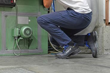 A person in blue pants and a white top is kneeling in front of a green machine. They are wearing black work shoes with blue laces. A fan is visible in the background.